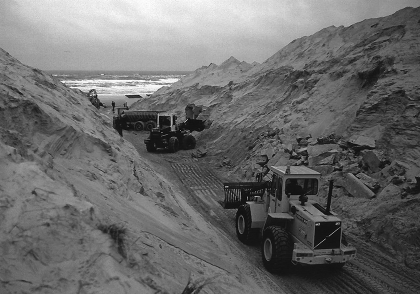 Zwartwit-foto met graafmachines in de duinen, de zee op de achtergrond